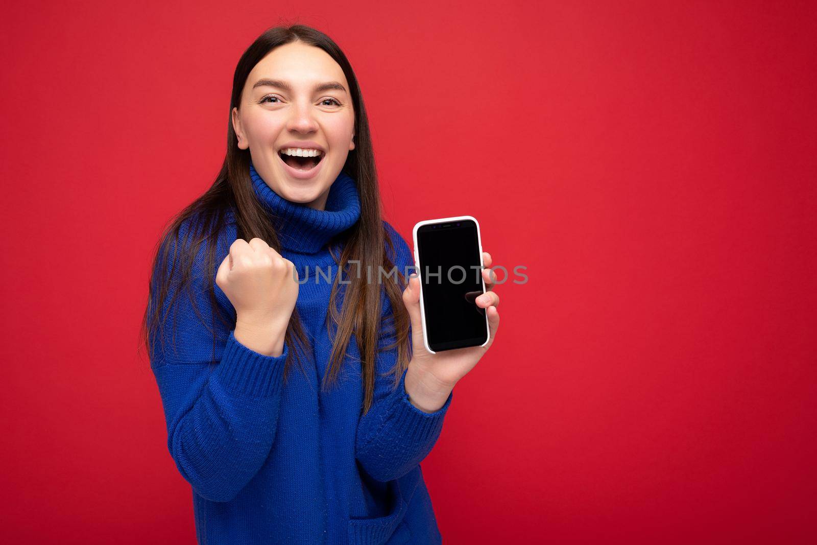 beautiful happy young brunette woman wearing casual blue sweater isolated over red background with empty space holding in hand mobile phone and showing smartphone with empty screen for mockup looking at camera.