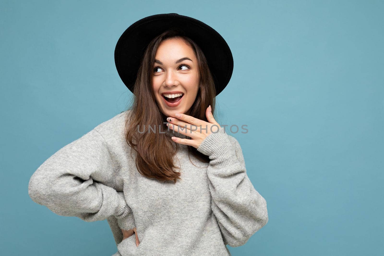 Portrait of young positive happy surprised beautiful brunette woman with sincere emotions wearing stylish grey pullover and black hat isolated over blue background with copy space.