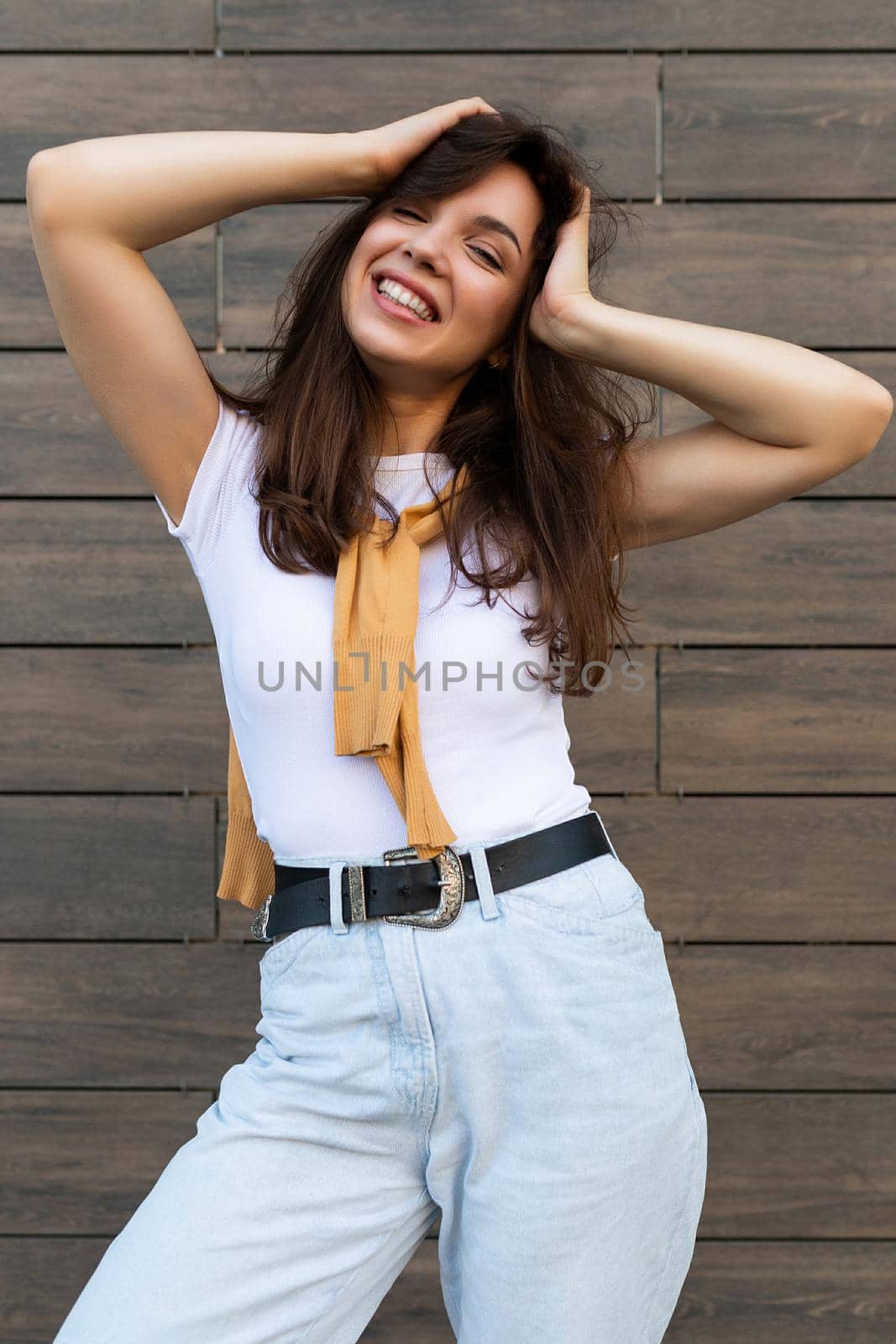 Vertical ptoto of young beautiful smiling positive happy brunette woman standing against brown wall in the street and wearing stylish outfit.