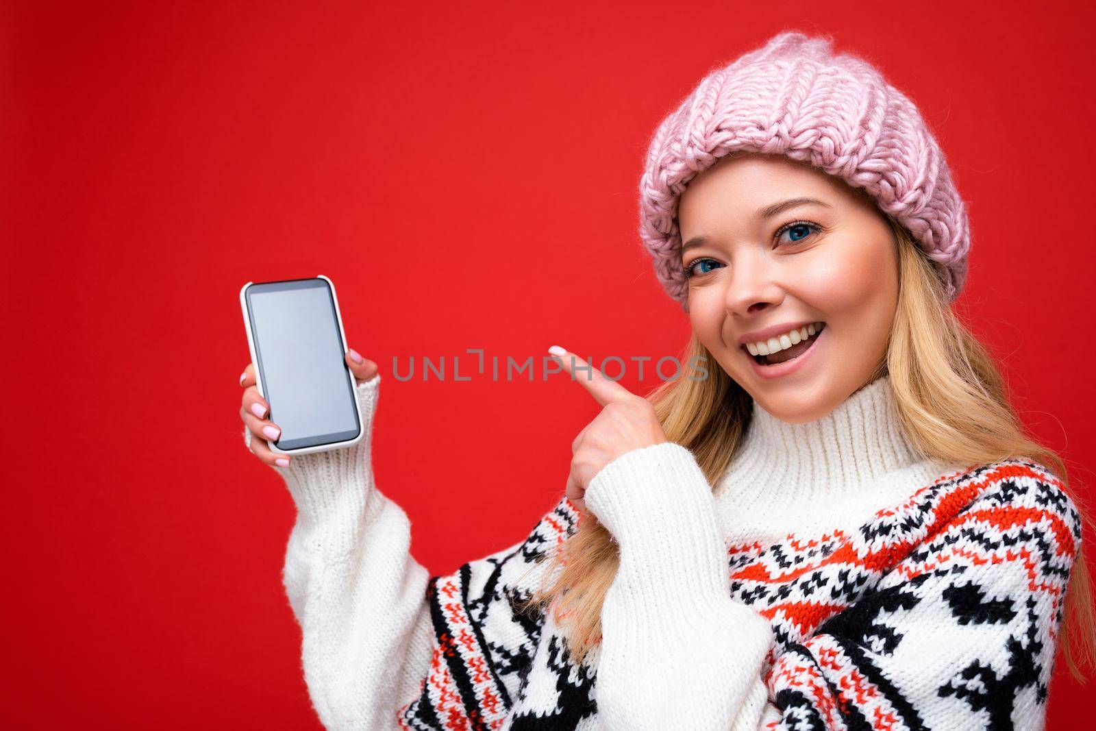 Photo of attractive smiling young blonde woman wearing warm knitted hat and winter warm sweater standing isolated over red background showing smartphone with empty screen for mock up looking at camera and pointing finger at gadjet.
