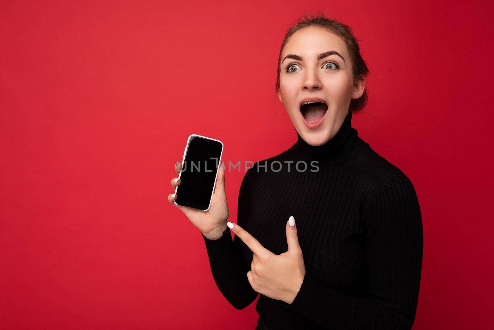 Photo of Shocked attractive positive young brunette woman wearing black sweater standing isolated over red background showing mobile phone with empty screen for mockup looking to the side pointing finger at gadjet.