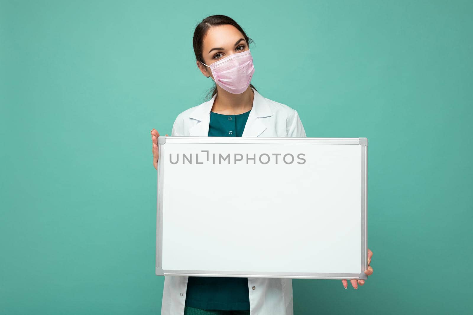 Woman doctor wearing a white medical coat and a mask holding blank board with copy space for text isolated on background. Coronavirus concept.
