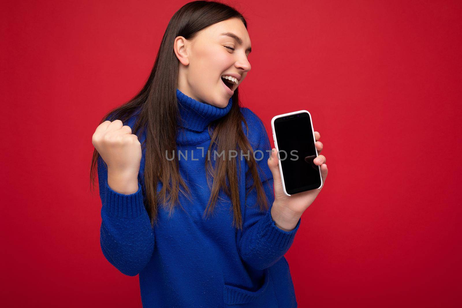 beautiful happy young brunette woman wearing casual blue sweater isolated over red background with empty space holding in hand mobile phone and showing smartphone with empty screen for mockup looking to the side by TRMK