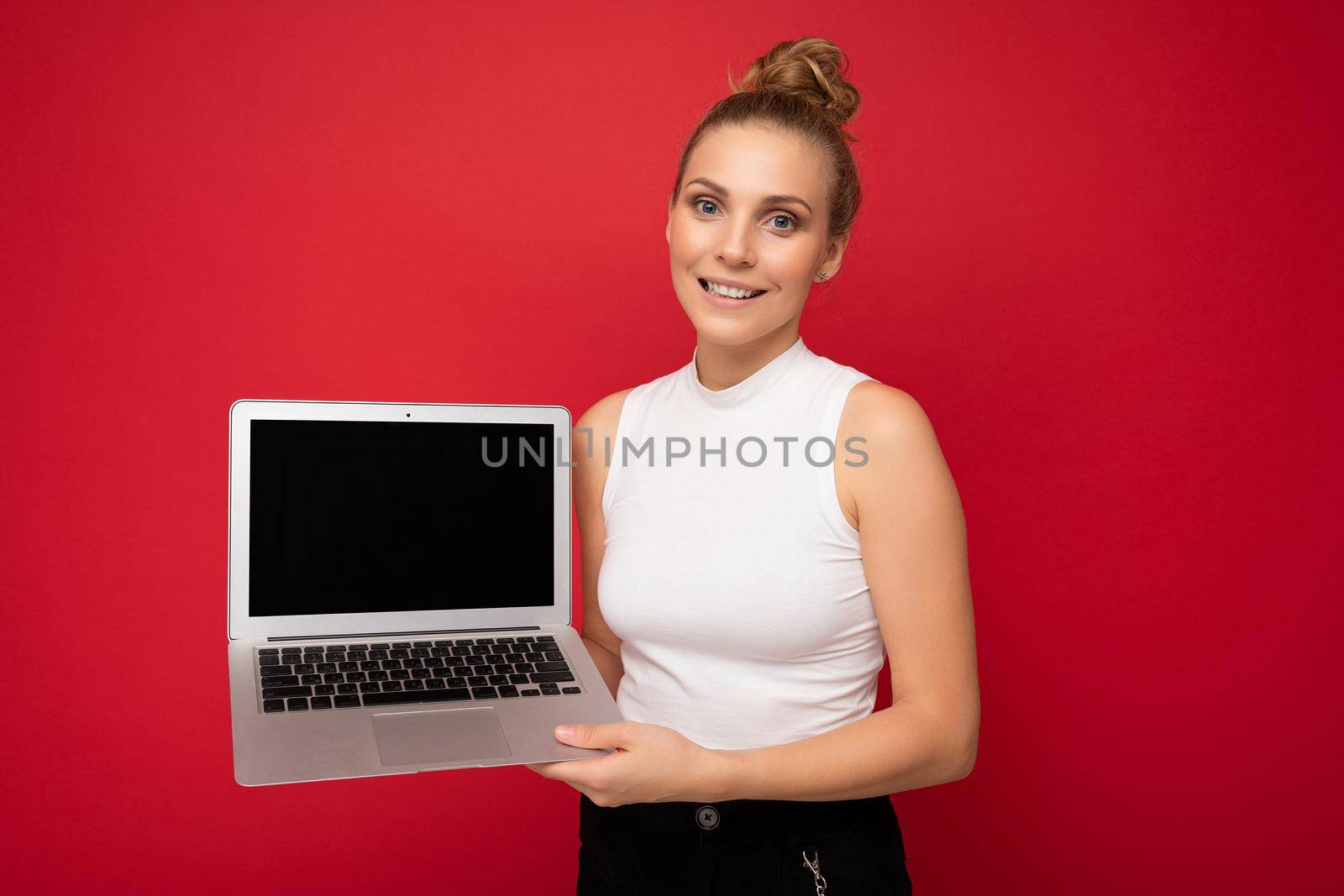 Photo of beautiful blond young woman with gathered hair looking at camera holding computer laptop with empty monitor screen with mock up and copy space wearing white t-shirt isolated over red wall background.
