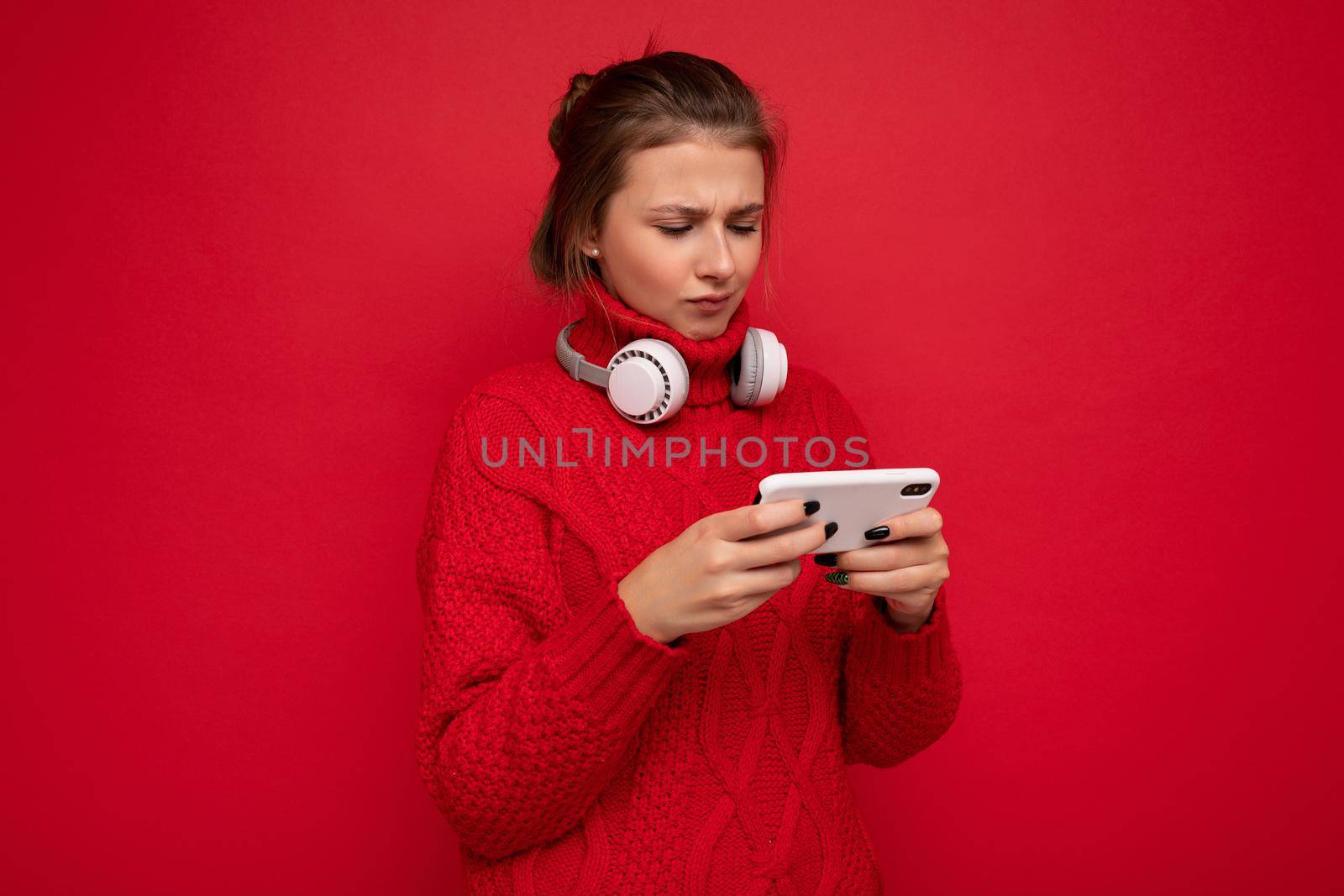 Attractive upset young brunet woman wearing red sweater isolated over red background wall holding and using mobile phone surfing on the internet and wearing bluetooth headphones looking at gadjet.