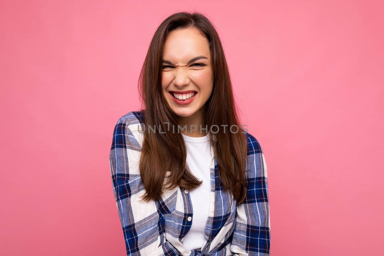 Photo portrait of young beautiful smiling hipster brunette woman in trendy blue and white shirt and jeans clothes. Sexy carefree female person posing isolated near pink wall with empty space in studio. Positive model with natural makeup by TRMK
