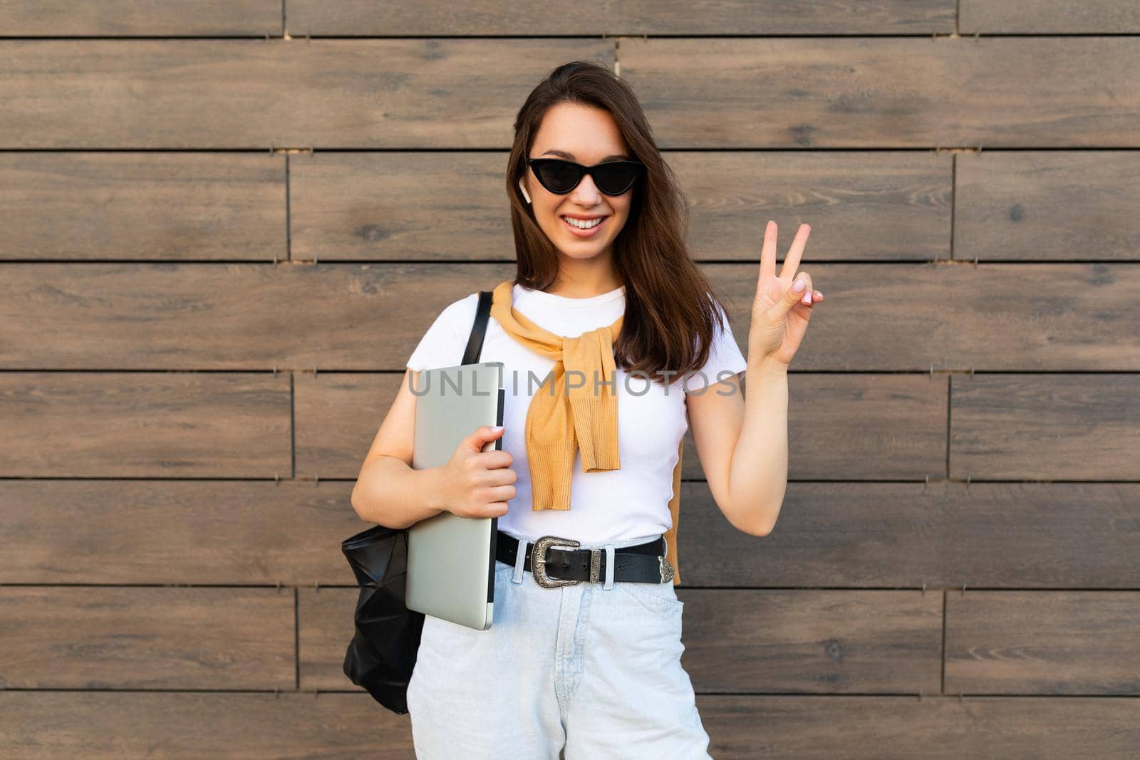 Beautiful smiling charming young brunet woman looking at camera holding computer laptop and sunglasses showing peace gesture in white t-shirt and light blue jeans in the street.