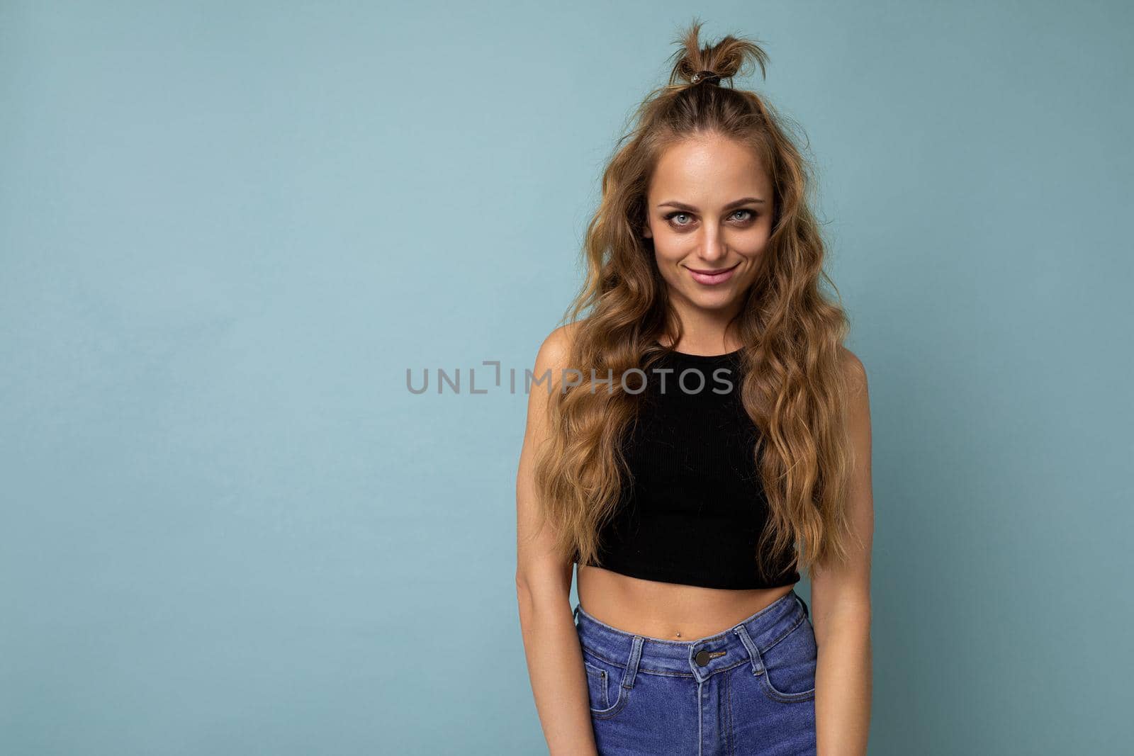 Young beautiful blond woman wearing black top t-shirt looking at camera. Positive female shows facial sincere emotions. Funny model isolated on blue wall with empty space by TRMK