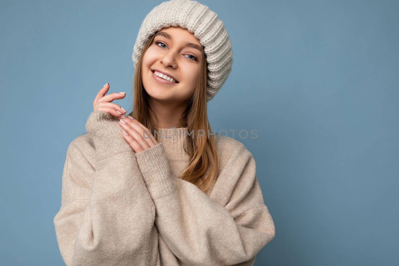 Portrait of fascinating smiling happy young dark blonde woman isolated over blue background wall wearing beige warm sweater and knitted beige hat looking at camera. Free space