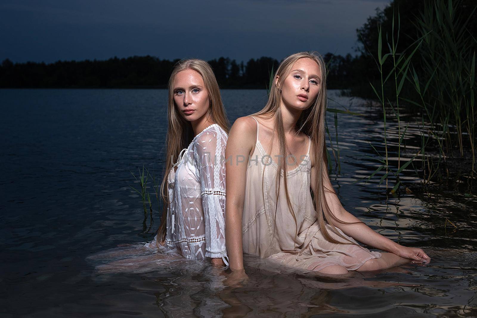 two young twin sisters posing in light dresses in water of lake at summer night by artemzatsepilin