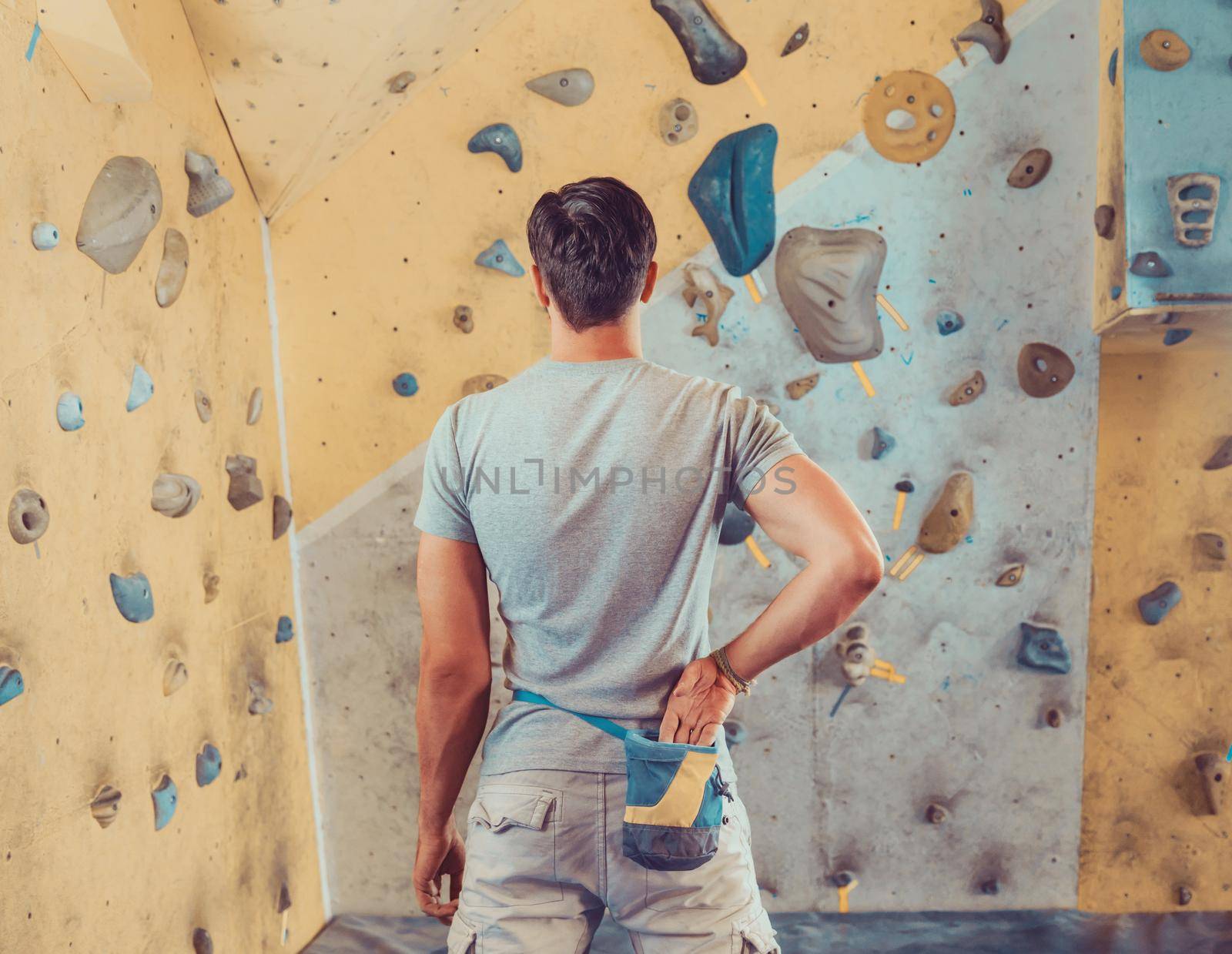 Sporty young man standing in front of a practical climbing wall indoor and preparing to climb. Climber putting his hand in the bag of powder magnesia