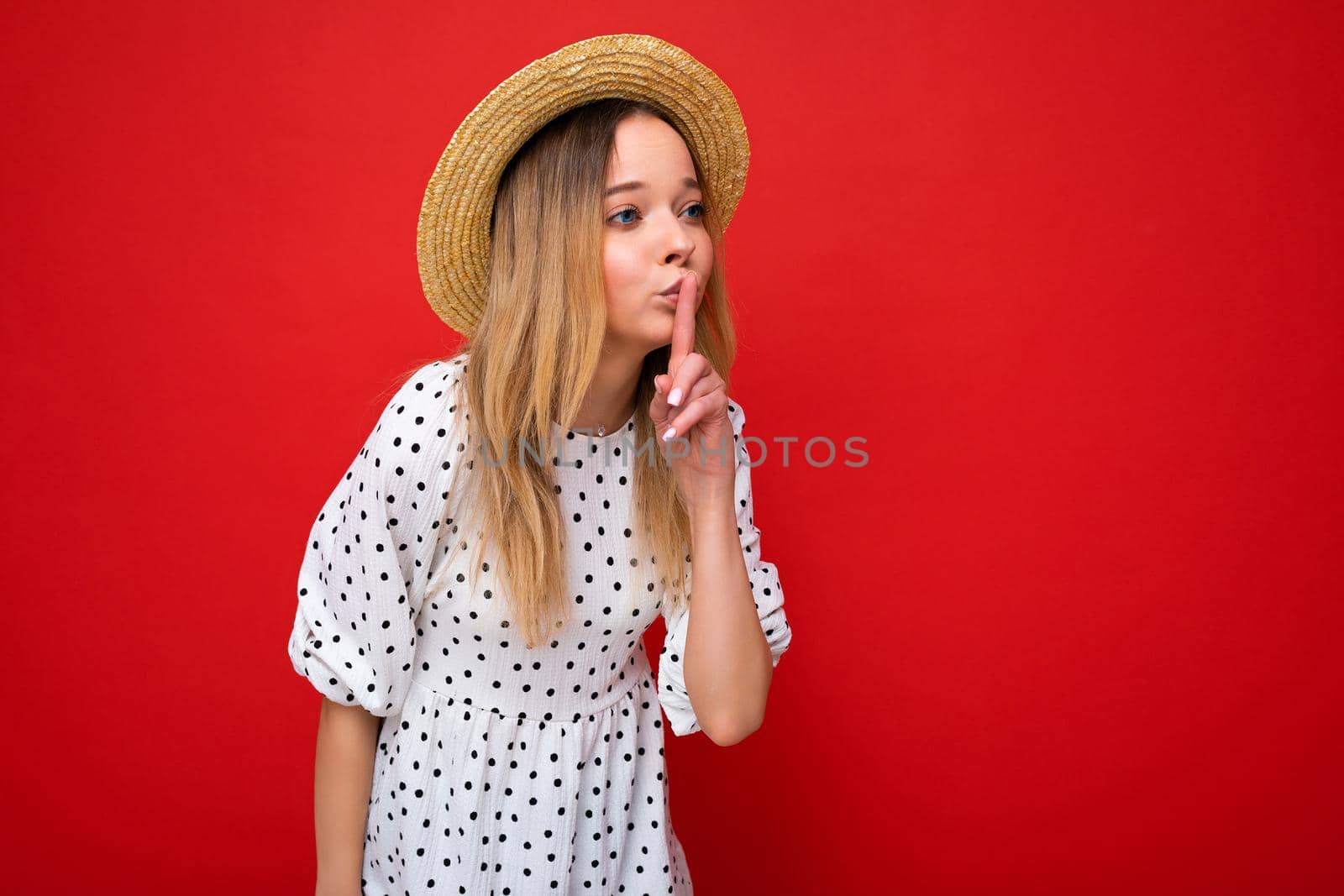Photo shot of young beautiful blonde woman wearing casual dress and straw hat isolated over red background with copy space showing shh geasture. Be quiet concept by TRMK