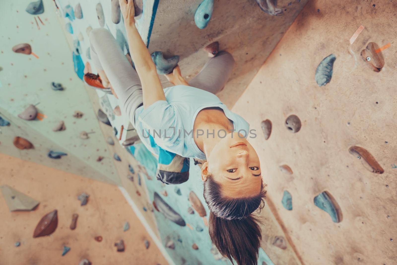 Smiling young woman climbing on practical wall indoor, bouldering, climber looking at camera