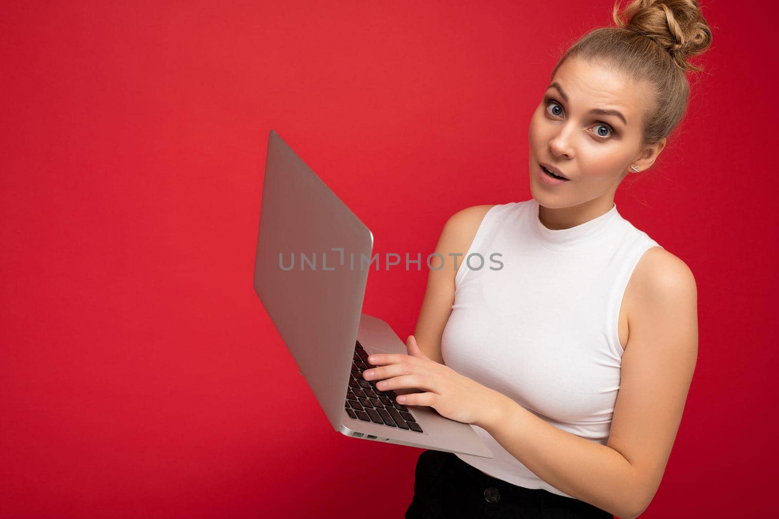 Side profile photo shot of beautiful asking amazed surprised blond young woman with gathered hair with open mouth wearing white t-shirt holding computer laptop typing on keyboard looking at camera isolated over red wall background.