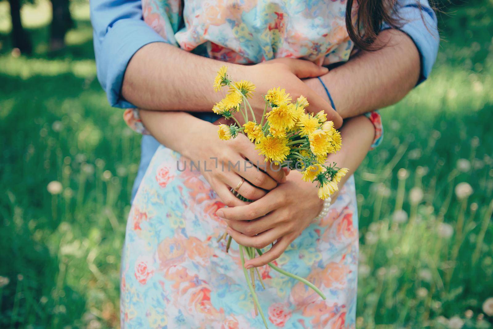 Married couple in love, man embraces a woman in summer park. Woman holding bouquet of yellow dandelions. Focus on flowers