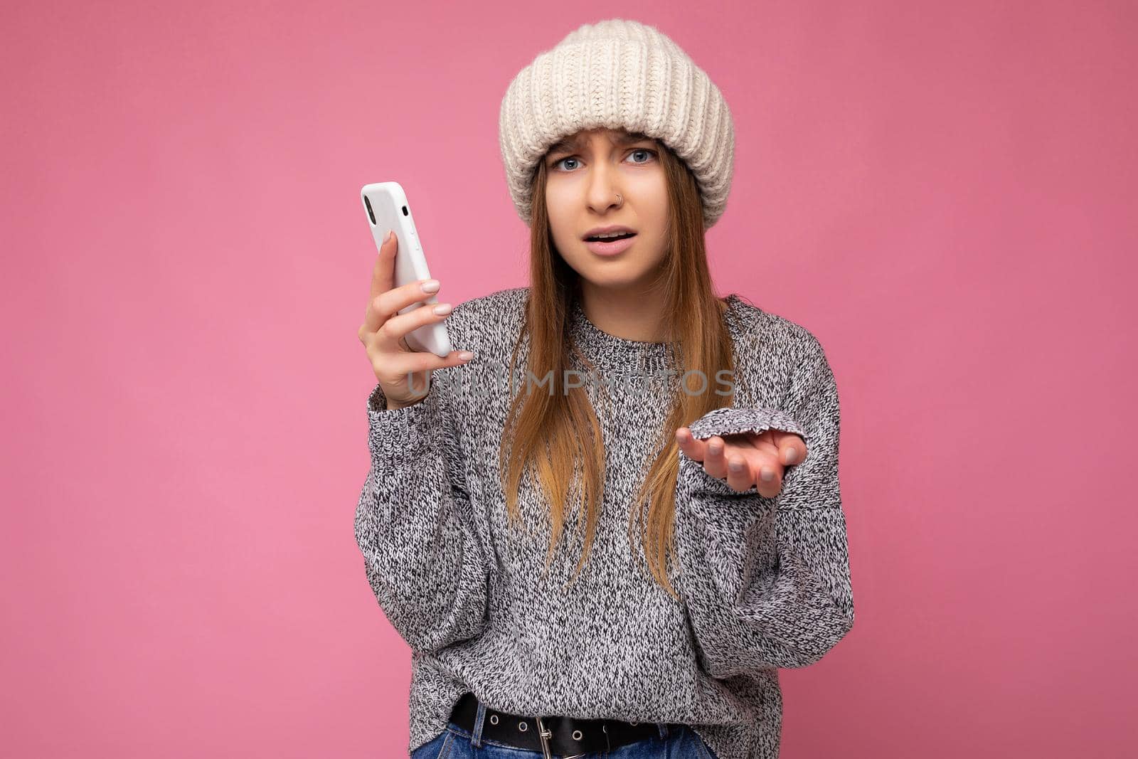 Beautiful angry upset young blonde woman wearing casual grey sweater and beige hat isolated over pink background holding in hand and using mobile phone looking at camera and don't understanding by TRMK