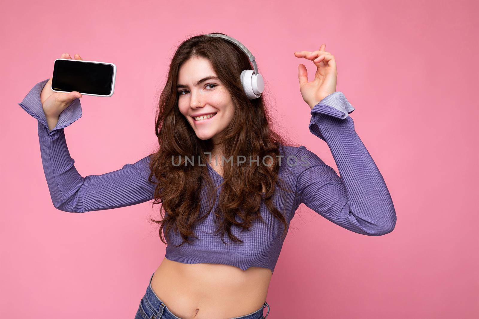 Beautiful happy smiling young woman wearing stylish casual outfit isolated on background wall holding and showing mobile phone with empty display for mockup wearing white bluetooth headphones listening to music and having fun looking at camera.