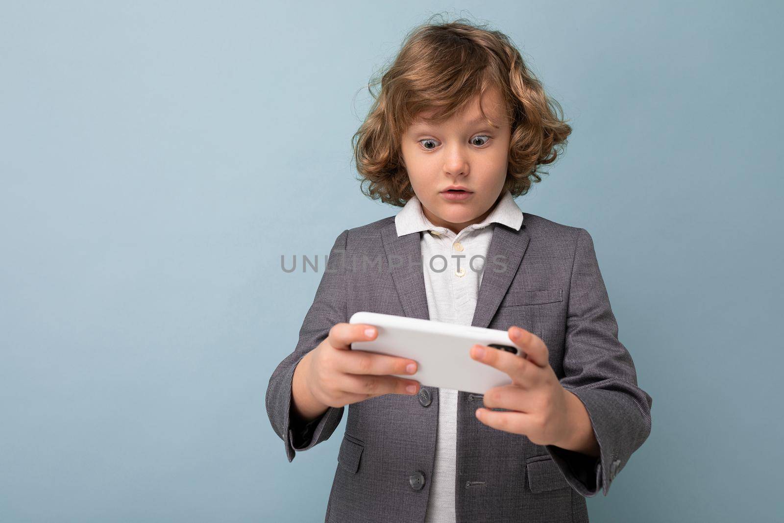 Photo shot of Handsome emotional child boy with curly hair wearing grey suit holding and using phone isolated over blue background looking at smartphone playing games by TRMK