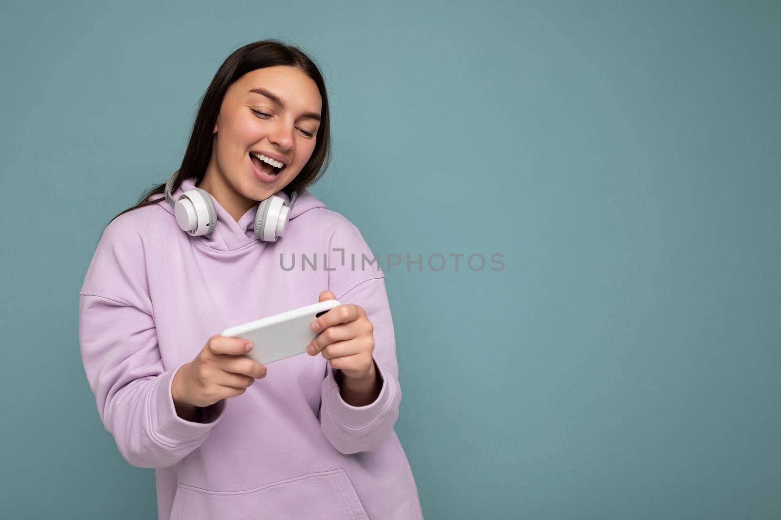 Photo shot of beautiful joyful smiling young female person wearing stylish casual outfit isolated over colorful background wall wearing white bluetooth wireless earphones and listening to music and using mobile phone looking at gadjet display by TRMK