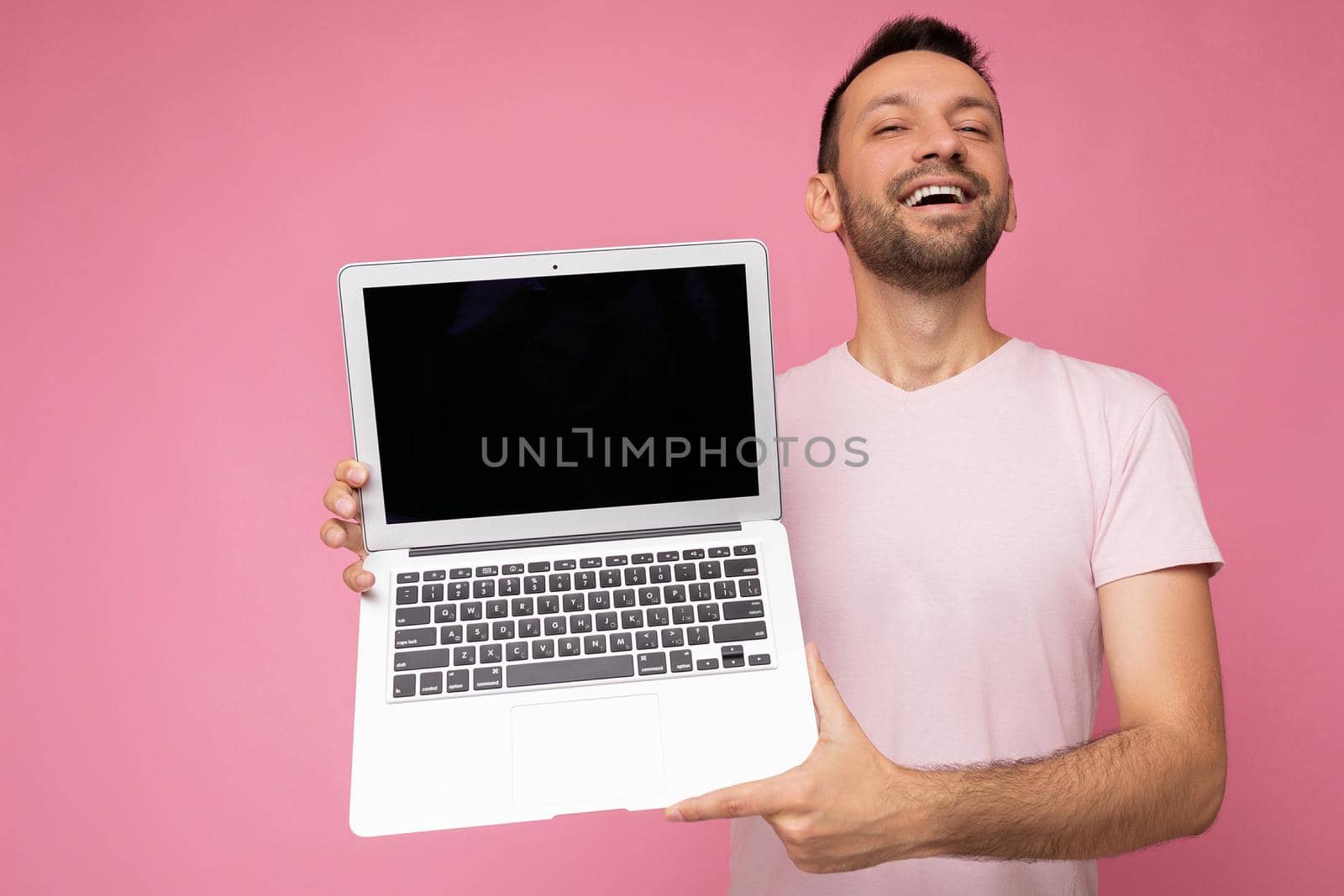 Handsome smiling brunet man holding laptop computer looking at camera in t-shirt on isolated pink background by TRMK