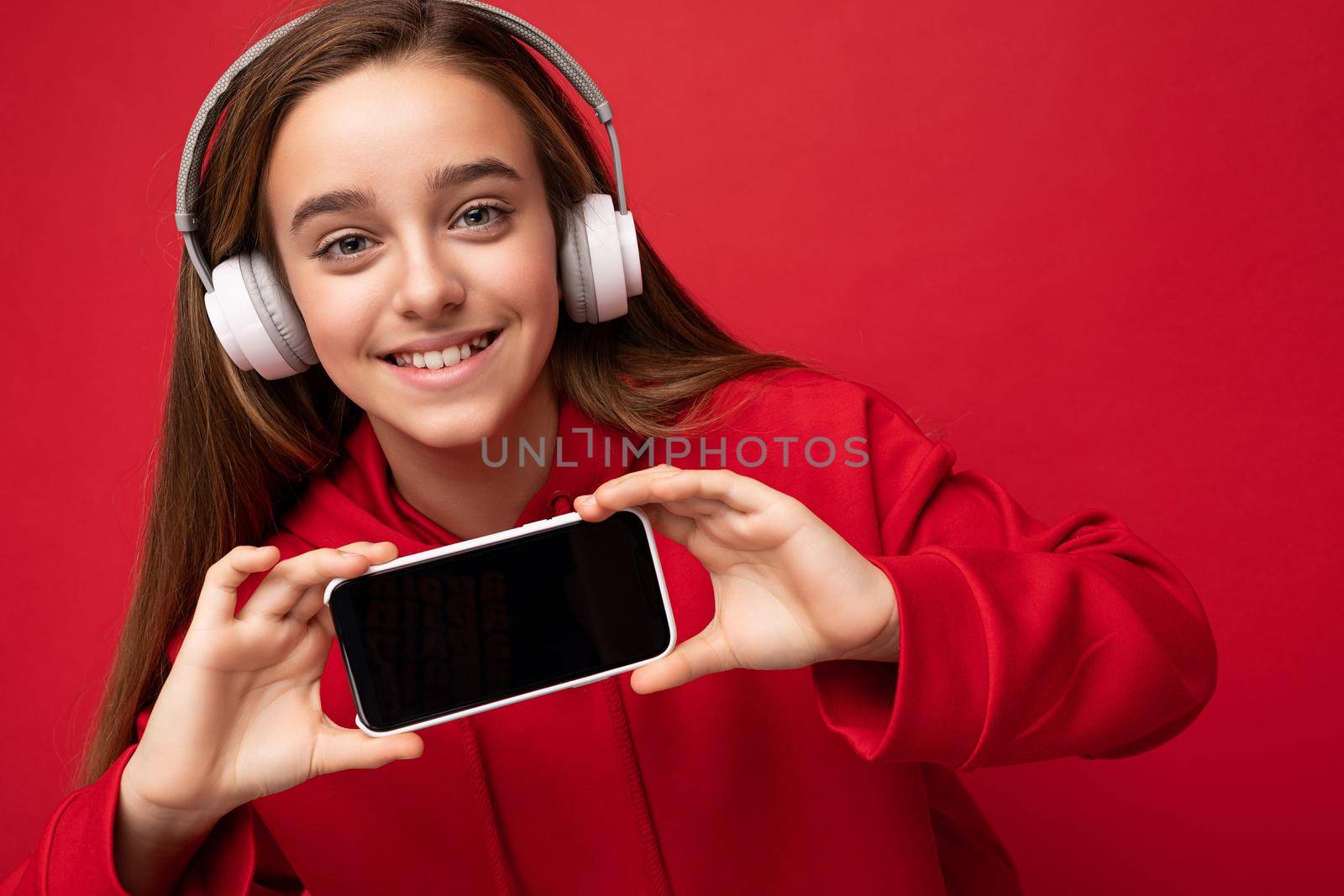 Closeup portrait of amazibg beautiful brunette girl wearing red hoodie isolated on red background holding and showing smartphone with empty display for cutout wearing white wireless headsets listening to energetic music looking at camera by TRMK