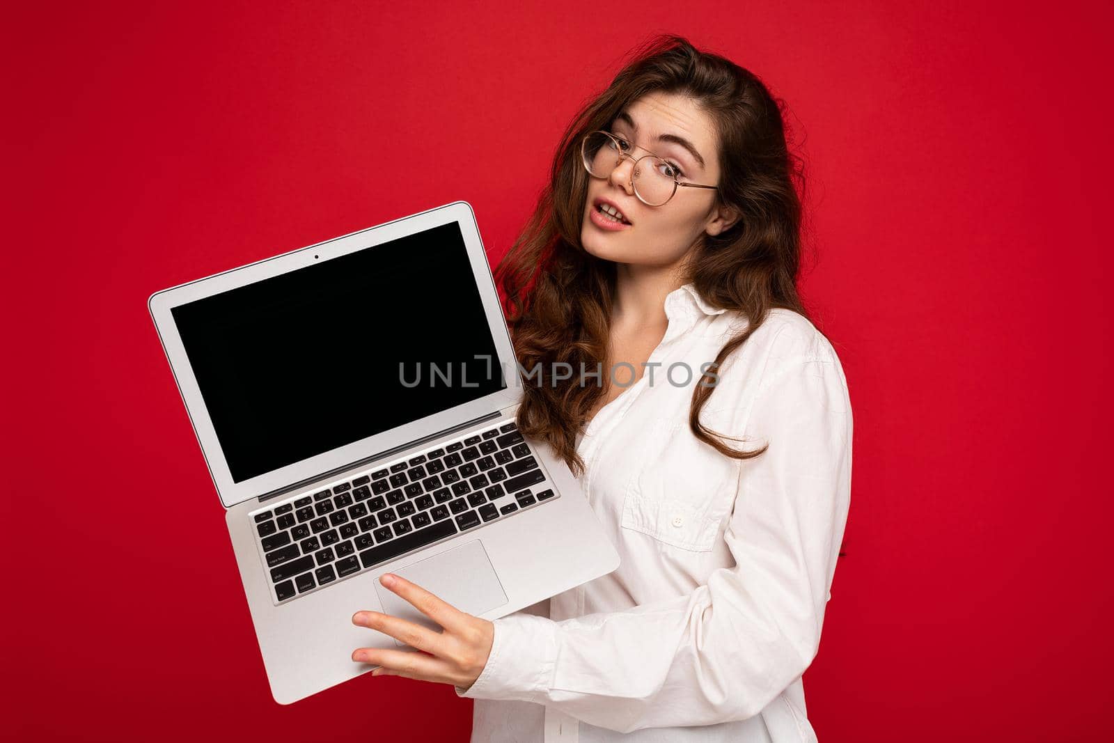 beautiful brunet curly young woman holding computer laptop wearing glasses white shirt looking at camera isolated over red wall background by TRMK