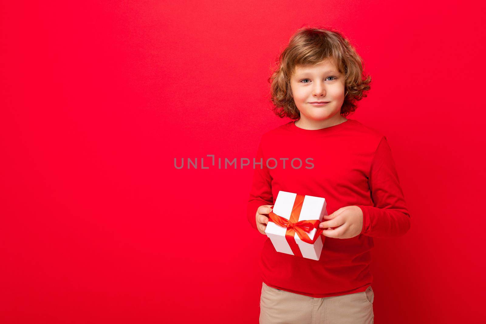 Shot of happy positive blond curly boy standing isolated over red background wall wearing red sweater holding gift box and looking at camera by TRMK