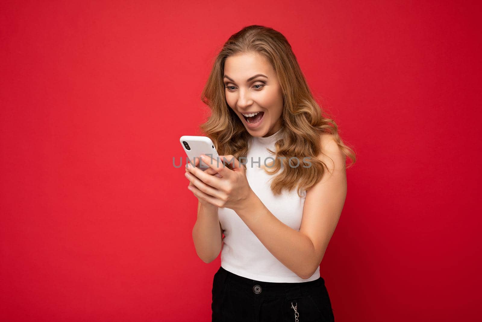 Photo shot of attractive surprised amazed positive good looking young woman wearing casual stylish outfit poising isolated on background with empty space holding in hand and using mobile phone messaging sms looking at smartphone display screen.
