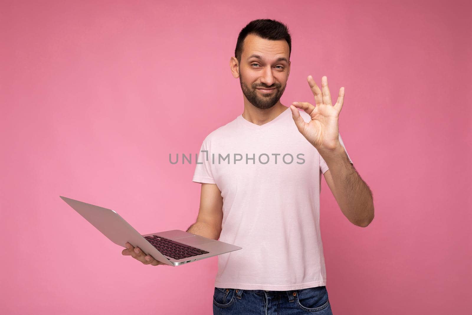 Handsome funny brunet man holding laptop computer and showing gesture okay looking at camera in t-shirt on isolated pink background.
