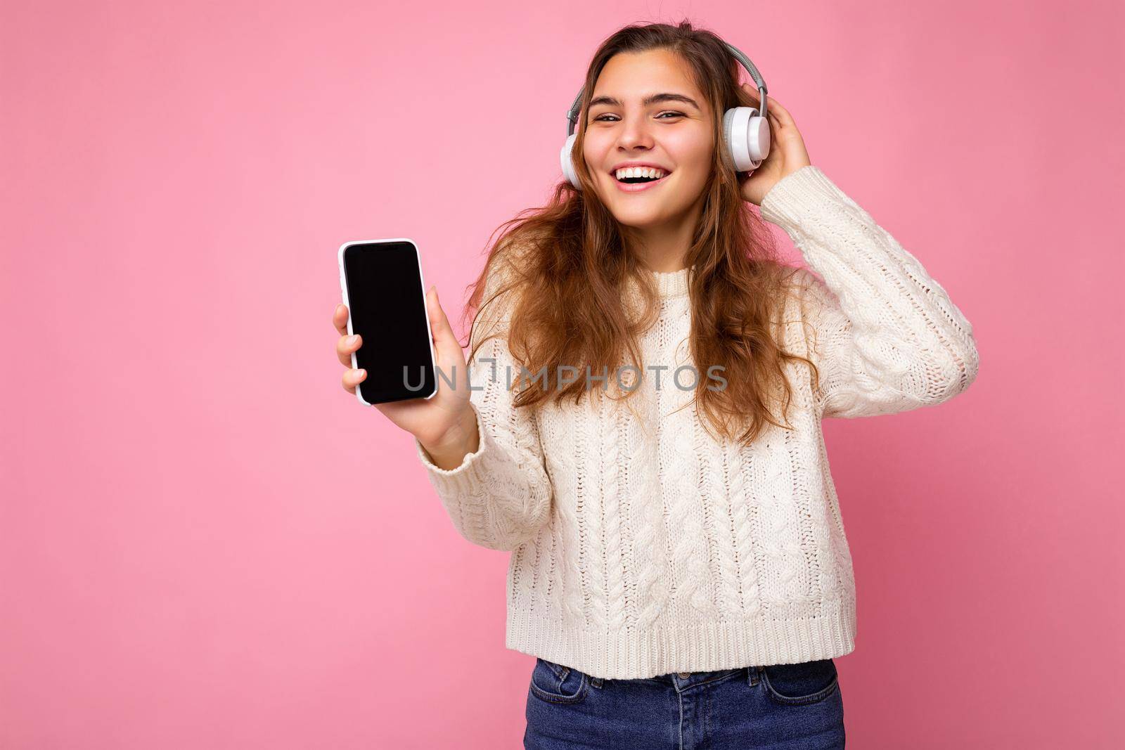 Beautiful happy smiling young woman wearing stylish casual outfit isolated on background wall holding and showing mobile phone with empty display for mockup wearing white bluetooth headphones listening to music and having fun looking at camera by TRMK