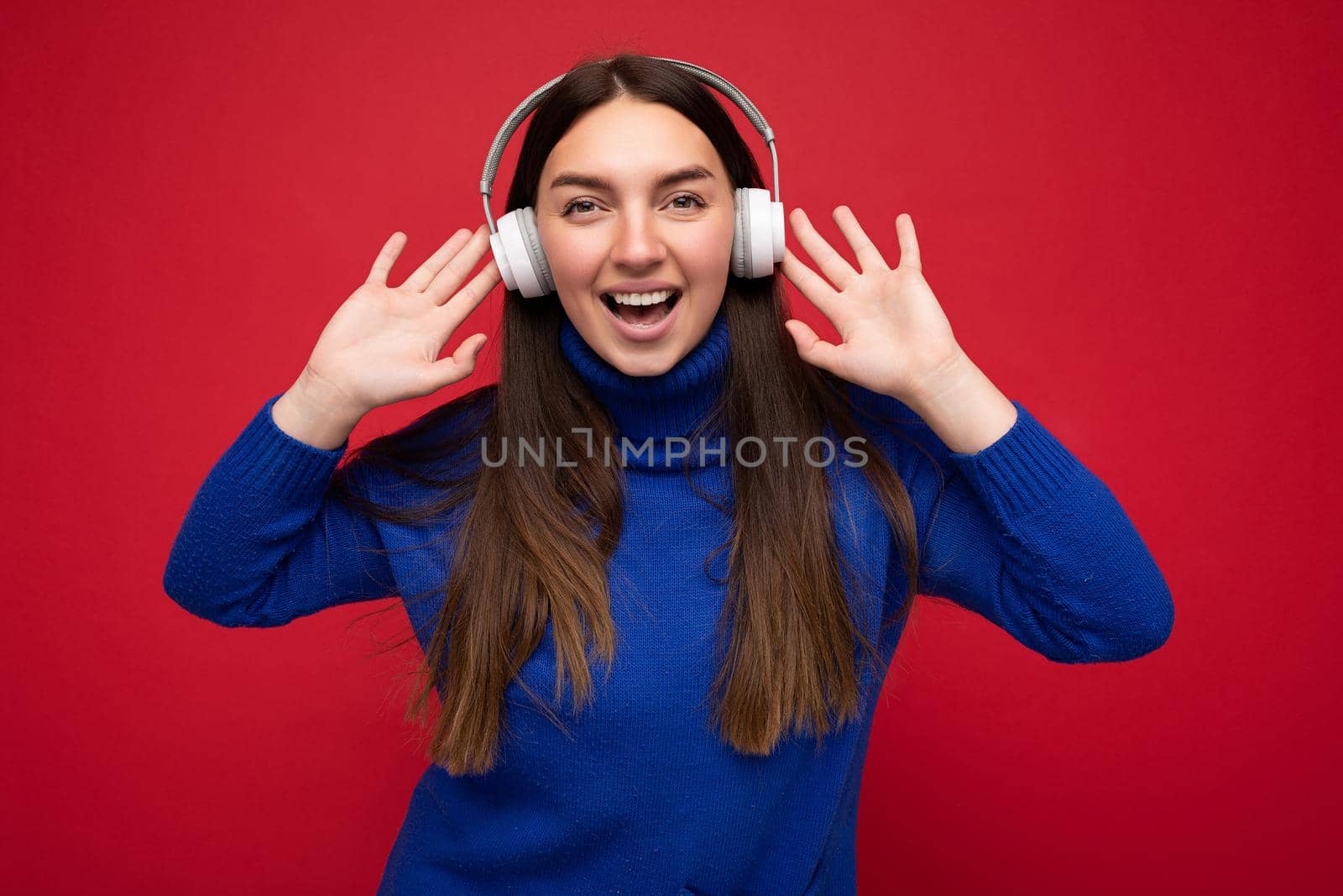 Attractive positive young brunette woman wearing blue sweater isolated over red background wall wearing white bluetooth earphones listening to cool music and having fun looking at camera.