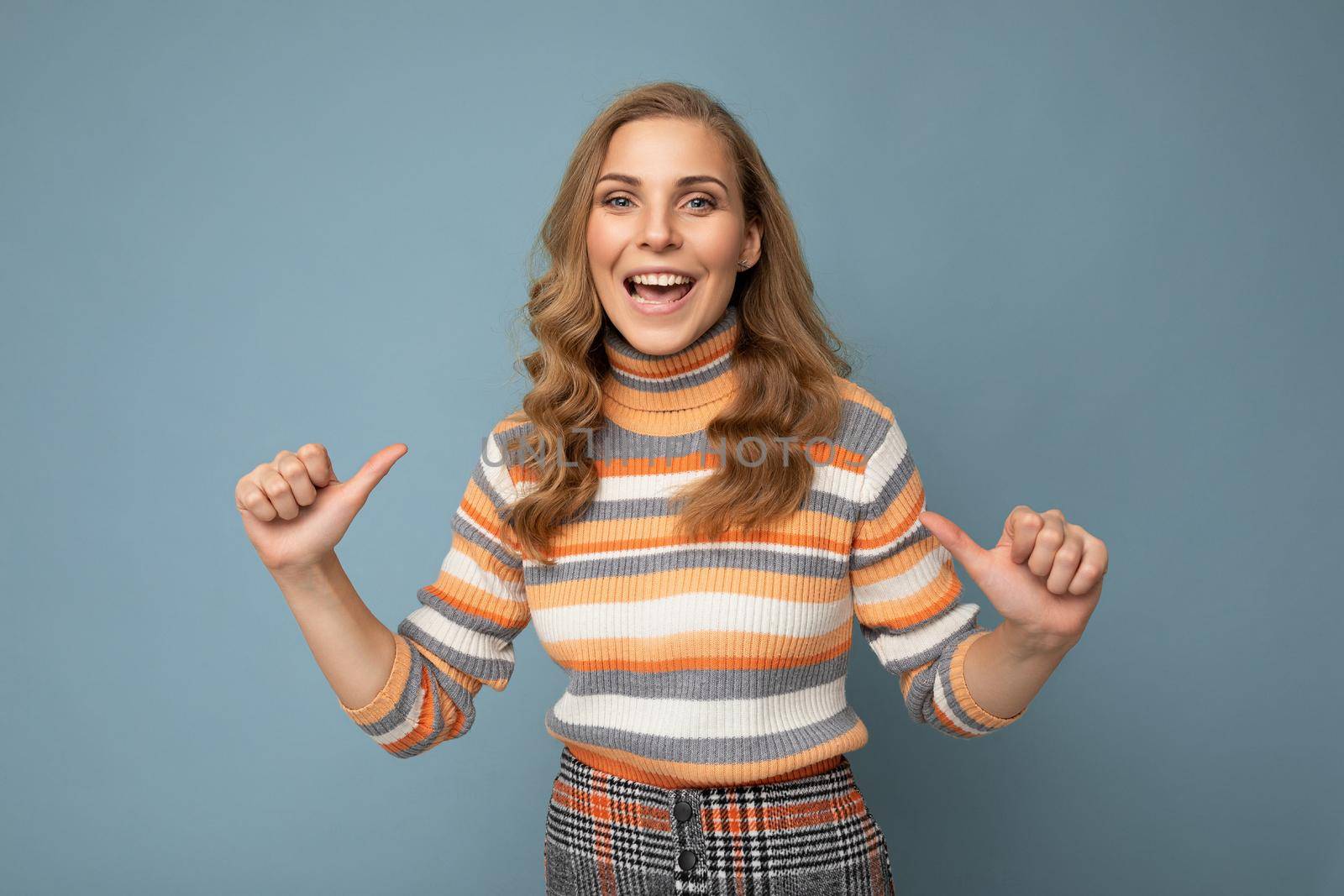 Portrait of young winsome attractive happy smiling blonde woman with wavy-hair wearing striped sweater isolated over blue background with empty space and pointing fingers at herself.