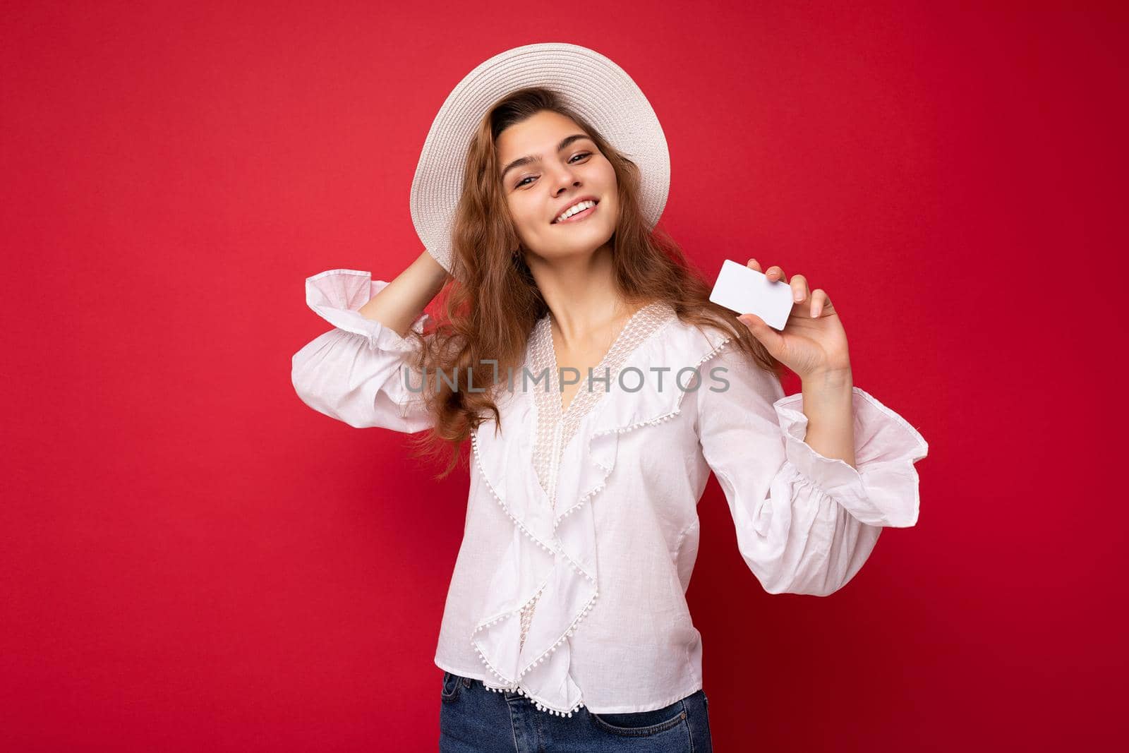 Shot of attractive positive smiling young dark blonde woman wearing white blouse and white hat isolated over red background holding credit card looking at camera by TRMK