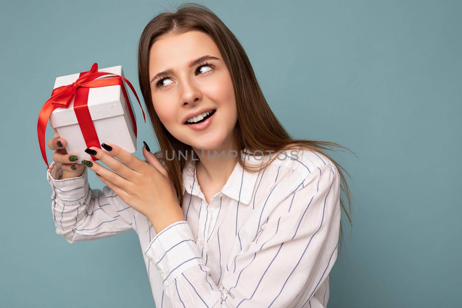 Photo shot of pretty positive surprised young dark blond woman isolated over colourful background wall wearing trendy outfit look holding gift box and looking at present box with red ribbon.