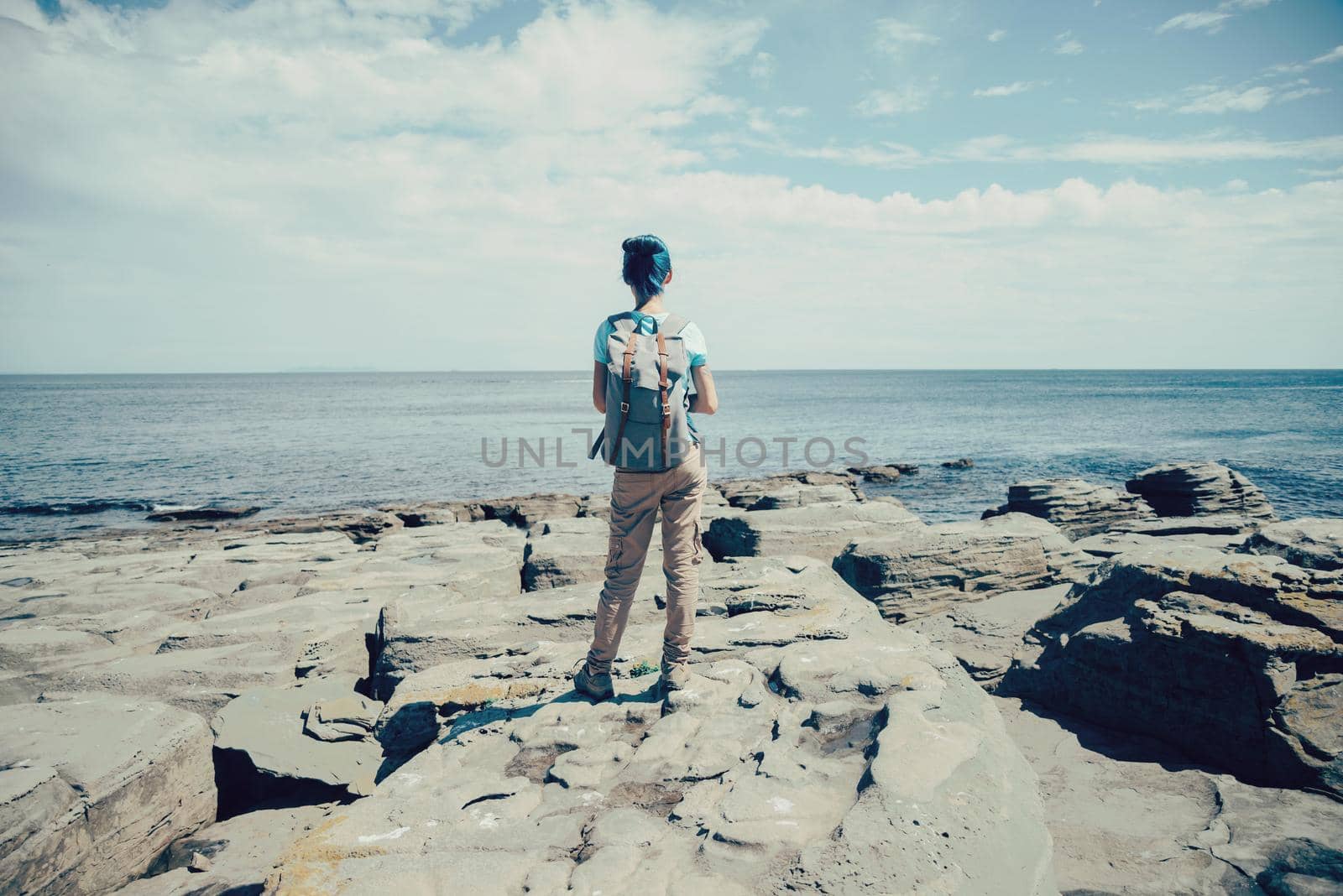 Traveler young woman standing on rocky coast and enjoying view of sea in summer, rear view