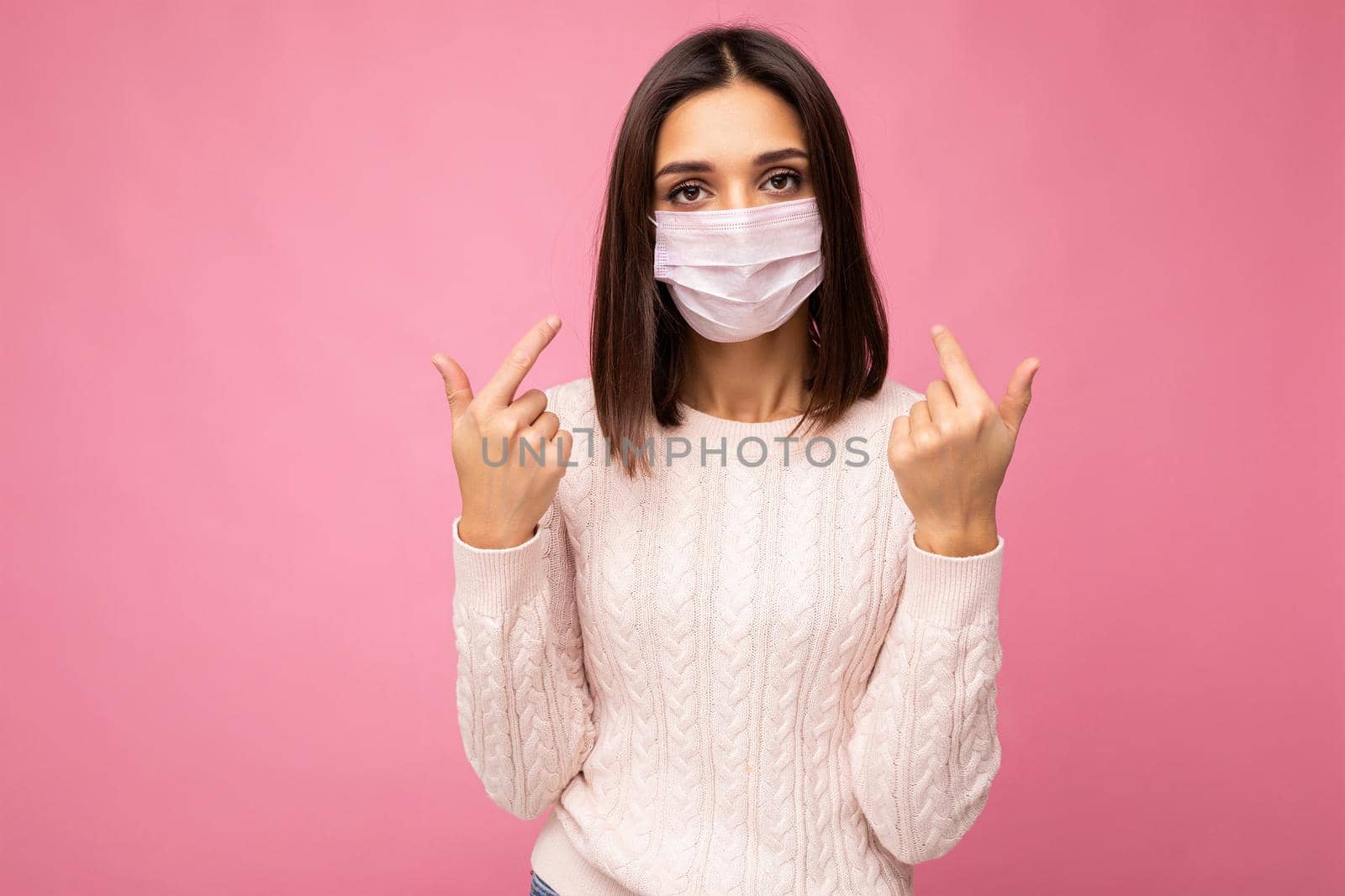 Young beautiful woman in reusable virus protective mask on face against coronavirus isolated on the pink background wall and pointing fingers at yourself by TRMK