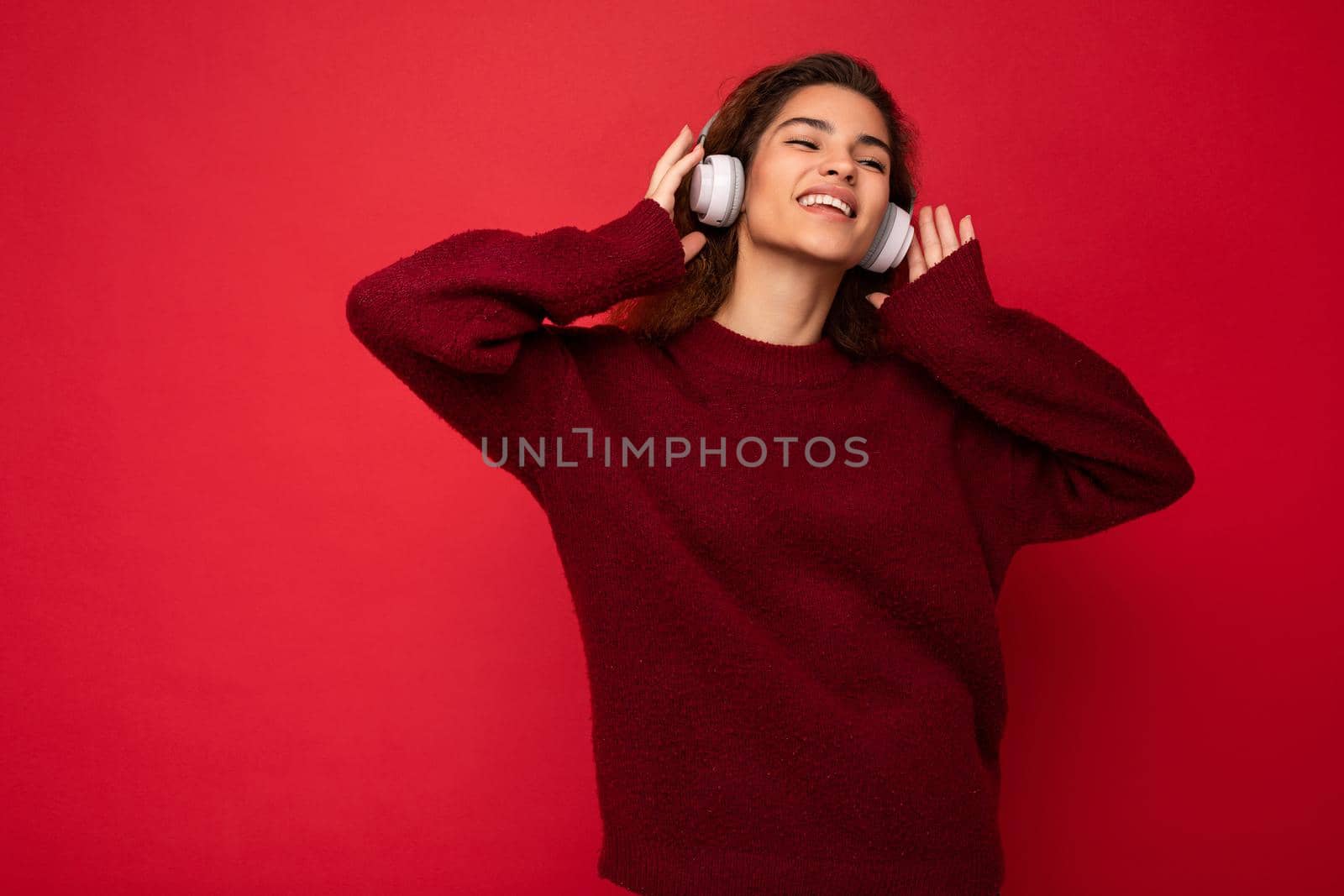 Photo of beautiful happy smiling young brunette curly woman wearing dark red sweater isolated over red background wall wearing white bluetooth headphones listening to music and having fun with close eyes by TRMK