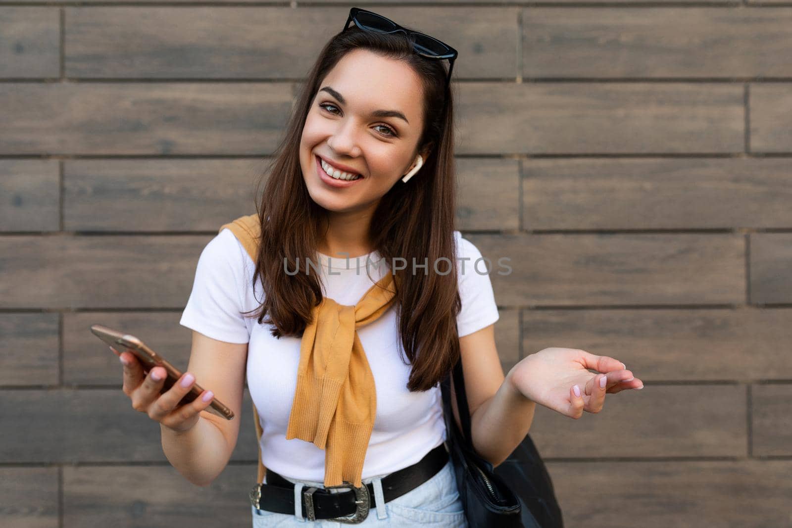 attractive happy smiling young woman wearing casual clothes standing in the street holding and using the mobile phone looking at camera.