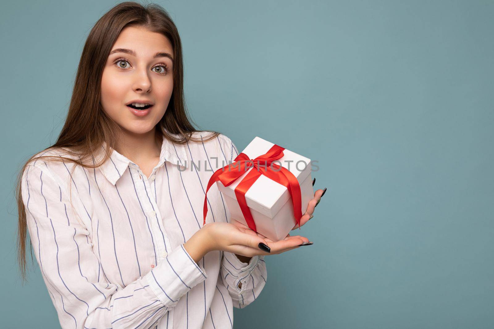 Photo of pretty positive surprised brunette young woman isolated over blue background wall wearing white shirt holding white gift box with red ribbon and looking at camera. Copy space, mockup