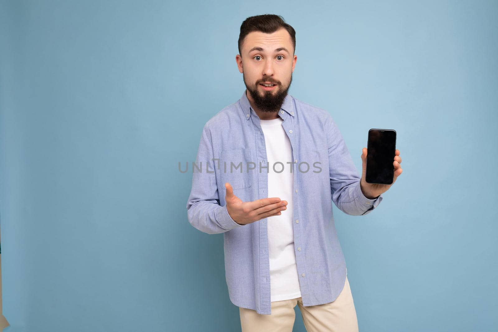 Photo of asking upset handsome good looking young brunette unshaven man with beard wearing casual white t-shirt and blue shirt poising isolated on blue background with empty space holding in hand mobile with empty screen looking at camera and pointing at gadjet by TRMK