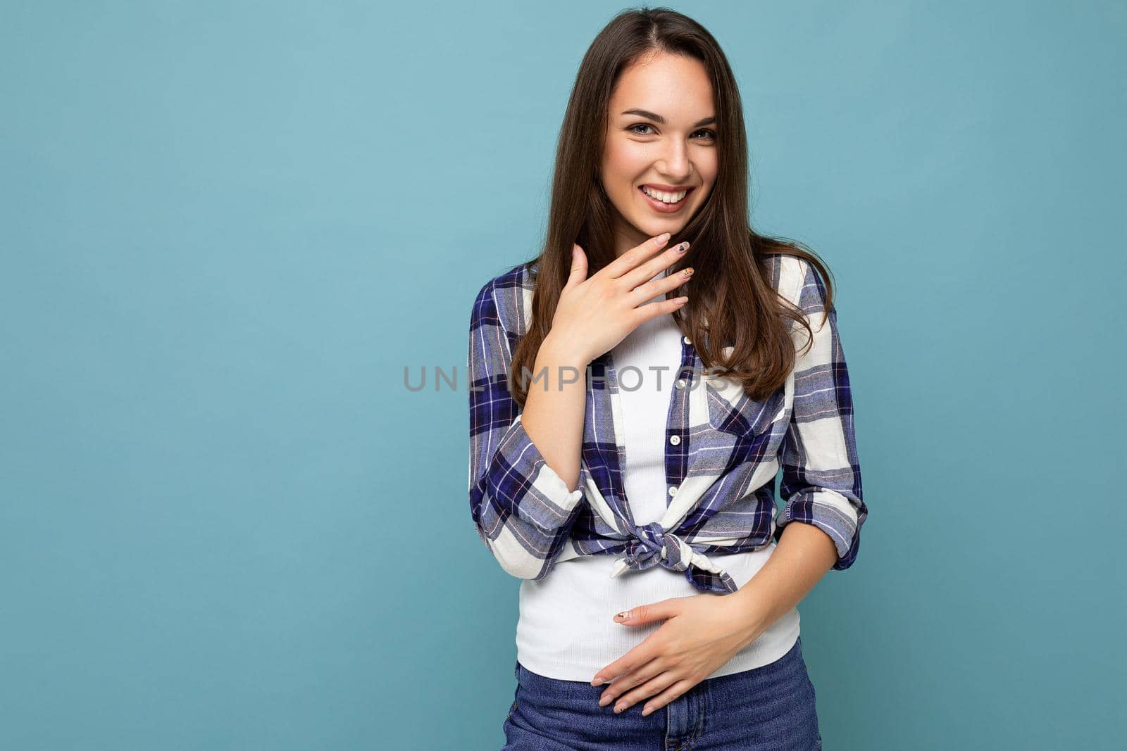 Young positive happy beautiful winsom brunette woman with sincere emotions wearing check shirt poising isolated over blue background with copy space and laughing.
