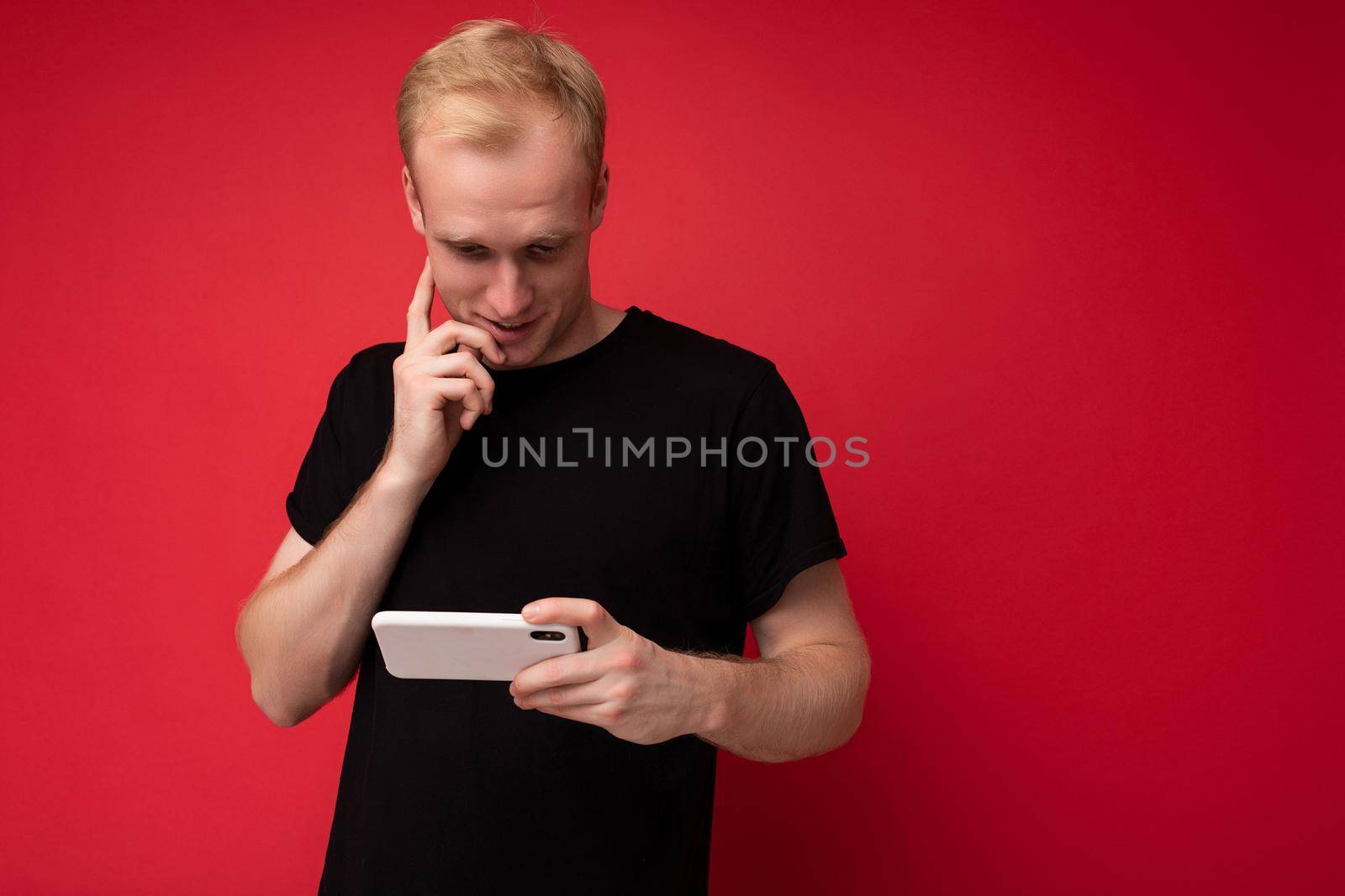 Photo of handsome positive young blonde man isolated over red background wall wearing black t-shirt holding and using mobile phone watching something via smartphone looking down at device screen.