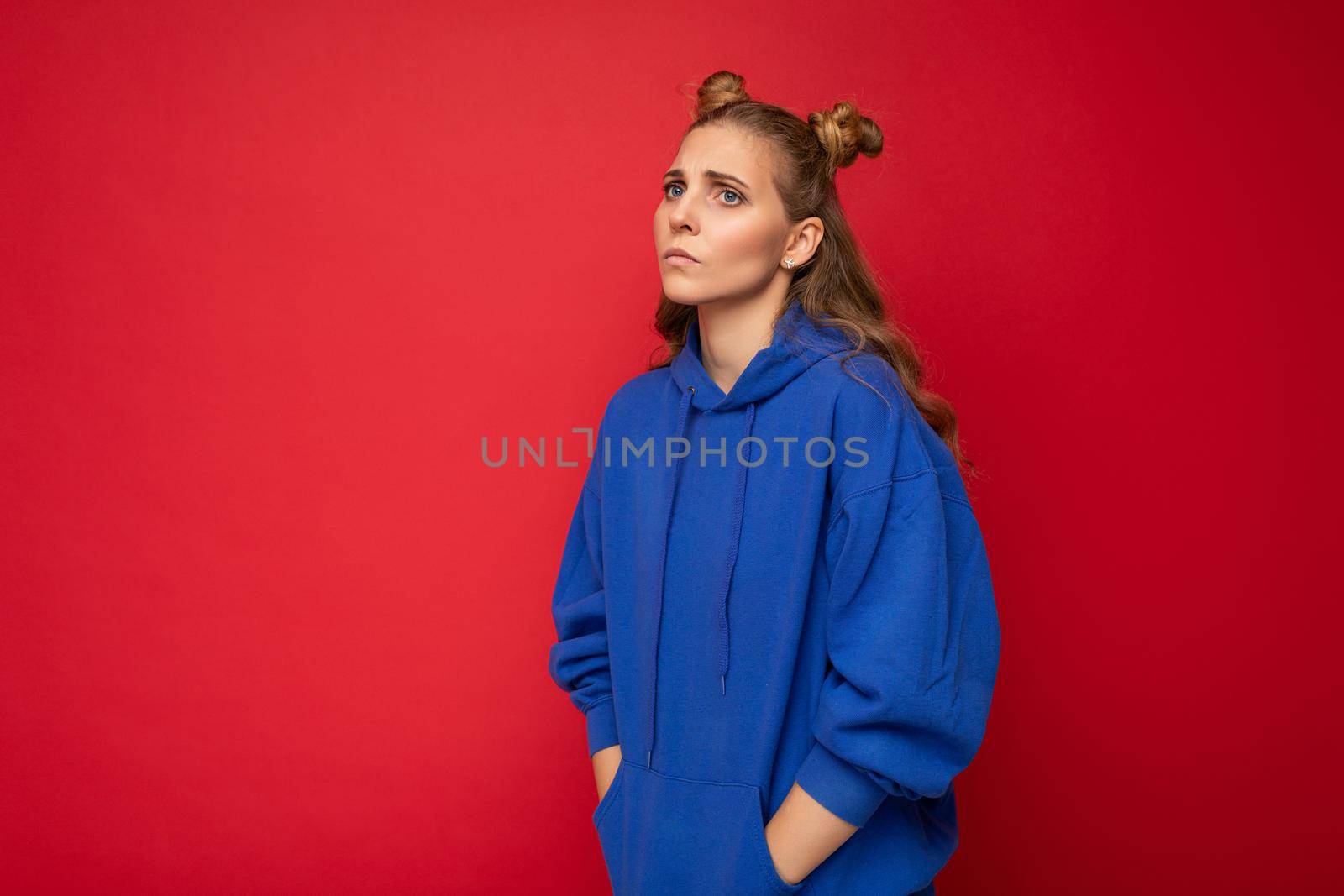 Portrait of sorrowful tired young attractive winsome blonde woman with two horns with sincere emotions wearing casual bright blue hoodie isolated on red background with free space.