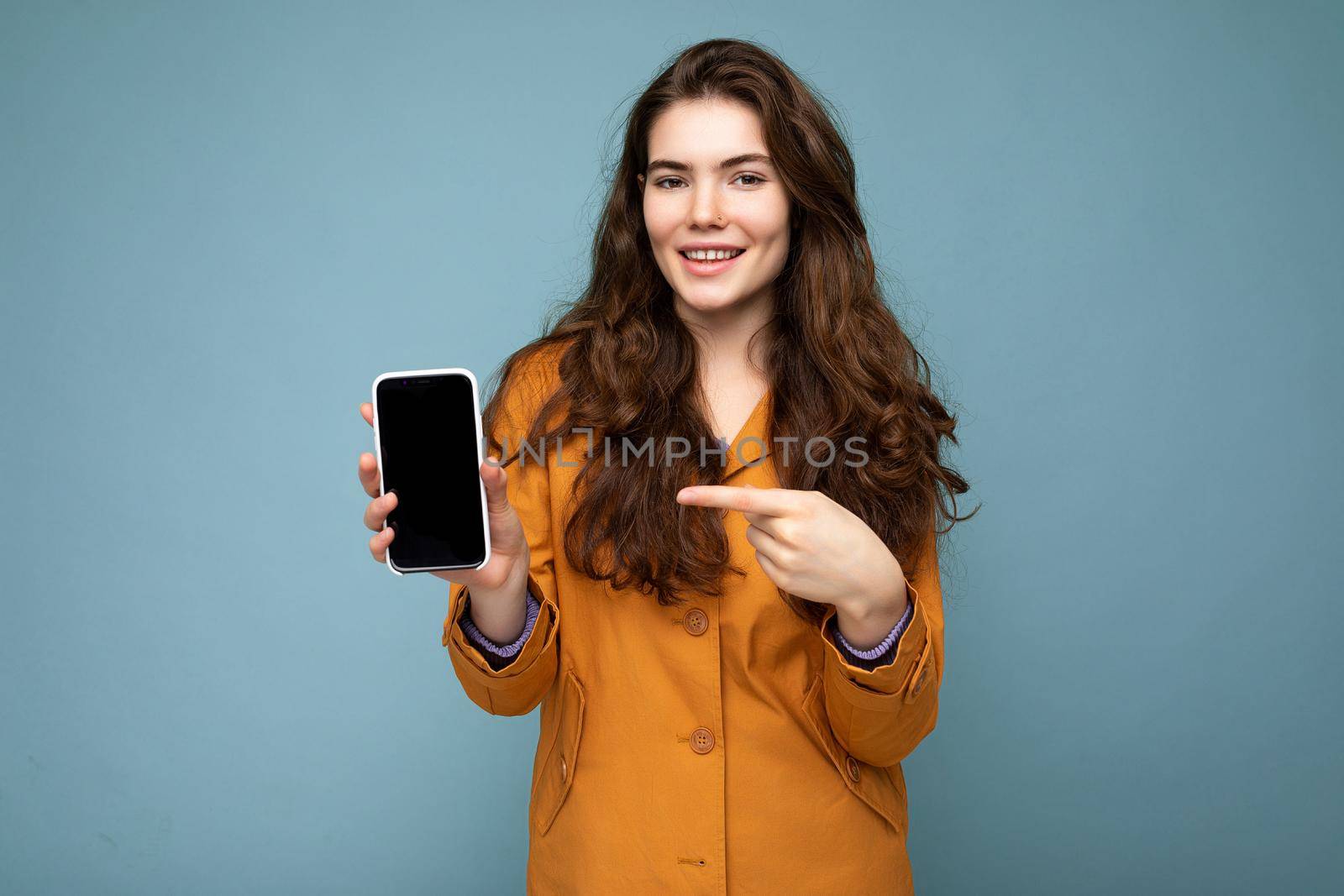 Photo of beautiful smiling young woman good looking wearing casual stylish outfit standing isolated on background with copy space holding smartphone showing phone in hand with empty screen display for mockup pointing at gadjet looking at camera by TRMK
