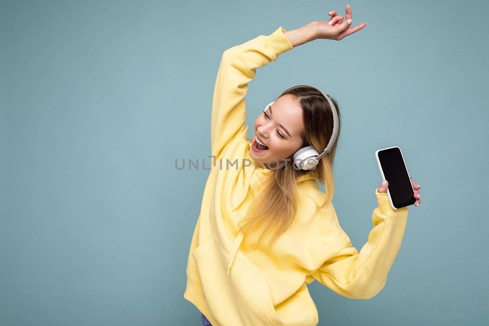 Photo of beautiful joyful smiling young woman wearing stylish casual clothes isolated over background wall holding and using mobile phone wearing white bluetooth headphones listening to music and having fun looking to the side by TRMK