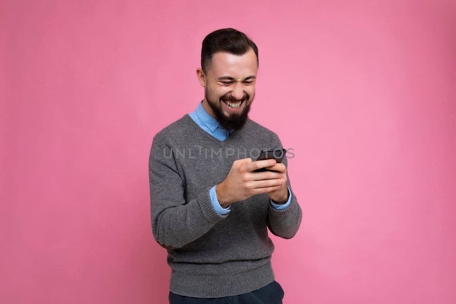 Photo shot of overjoyed cool good looking brunet unshaven young man with beard wearing grey sweater and blue shirt isolated over pink background wall with empty space holding in hand and using mobile phone messaging sms online looking down at telephone.