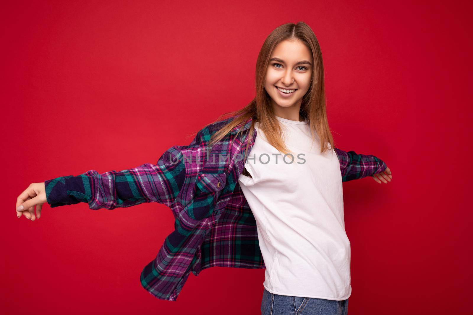 Photo of beautiful happy smiling young dark blonde woman isolated over red background wearing colourful stylish shirt and casual white t-shirt looking at camera and having fun by TRMK