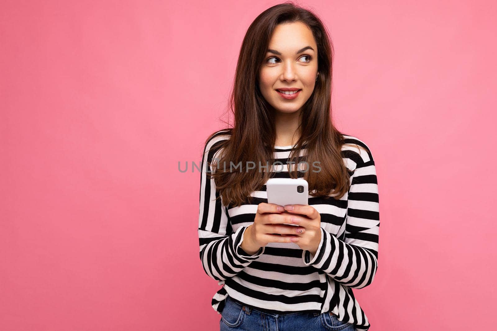 shot of pretty young smiling brunette woman using mobile phone communicating via texting sms wearing sweater isolated on wall background looking to the side by TRMK