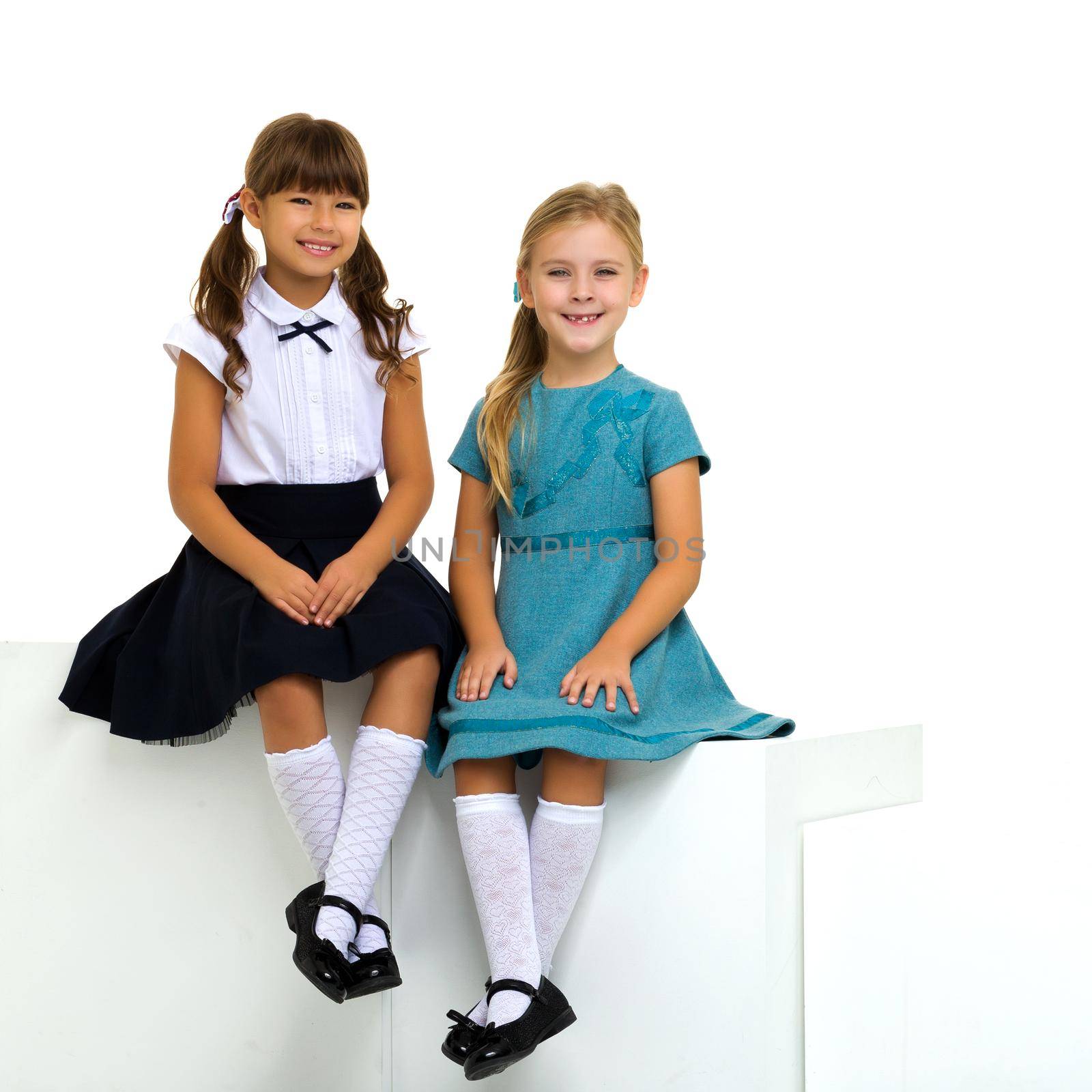 Full length shot of cute sitting girls. Above view of adorable sisters posing against white background. One of girls in stylish blue dress, the other in school uniform. Elementary school students