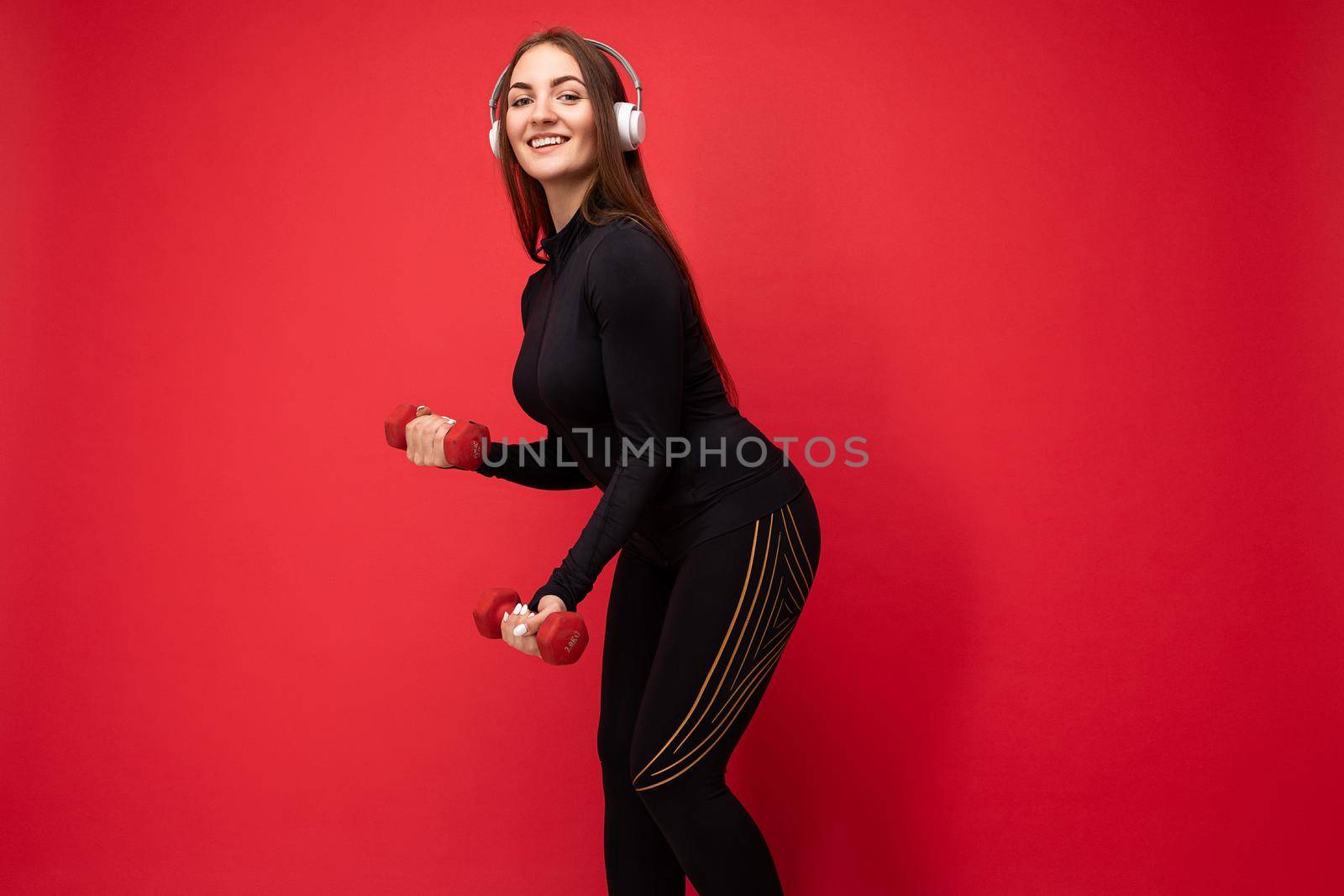 Side-profile photo of attractive positive smiling young brunette woman wearing black sport clothes isolated on red background wall doing fitness using dumbbells wearing white bluetooth earphones listening to good music looking at camera.